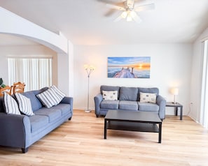A living room with two sofas, a coffee table, a floor lamp, a ceiling fan, and a sunset painting on the wall.