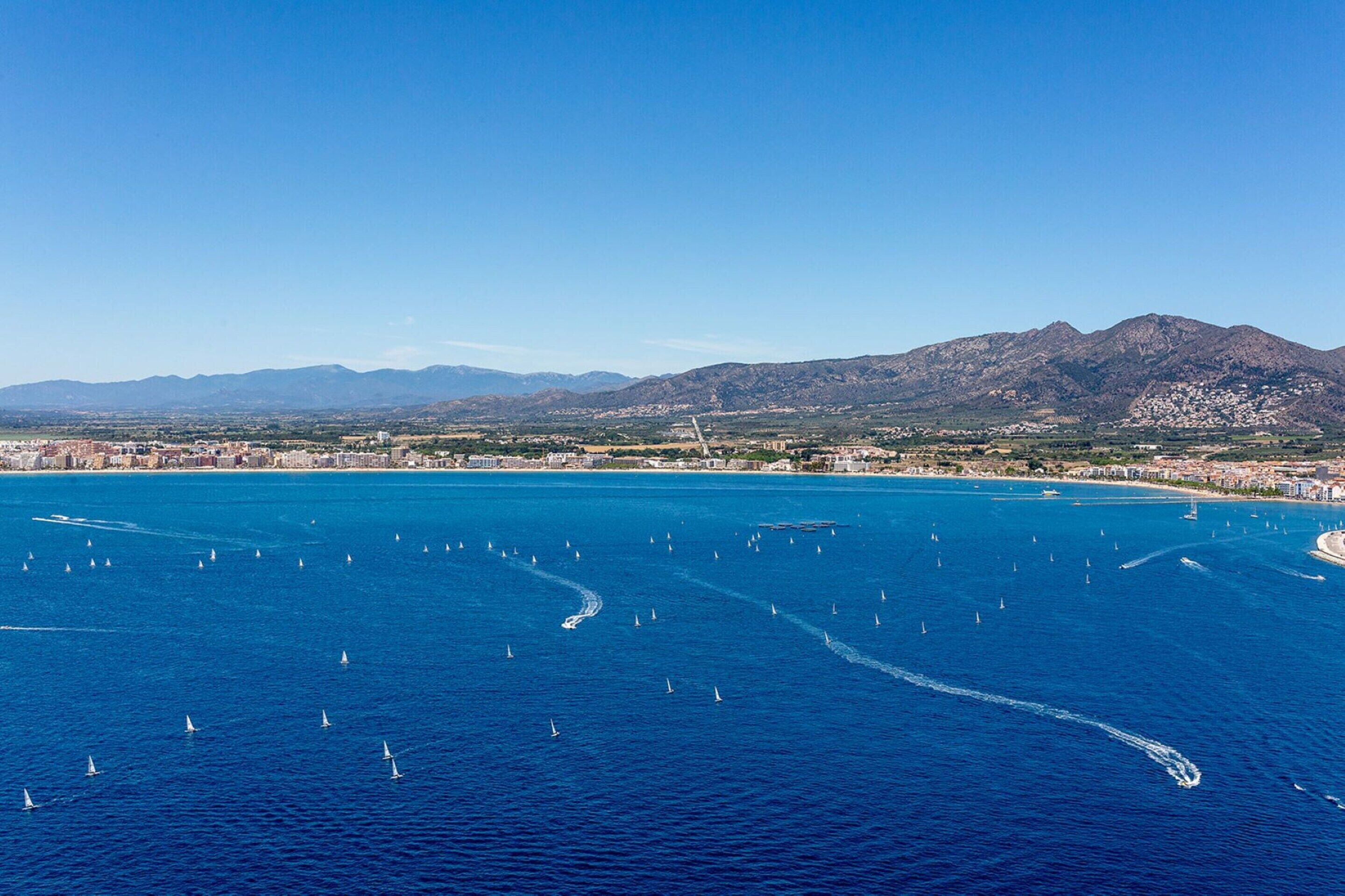 A coastal city with a clear blue sea, numerous sailboats, and a mountainous backdrop.