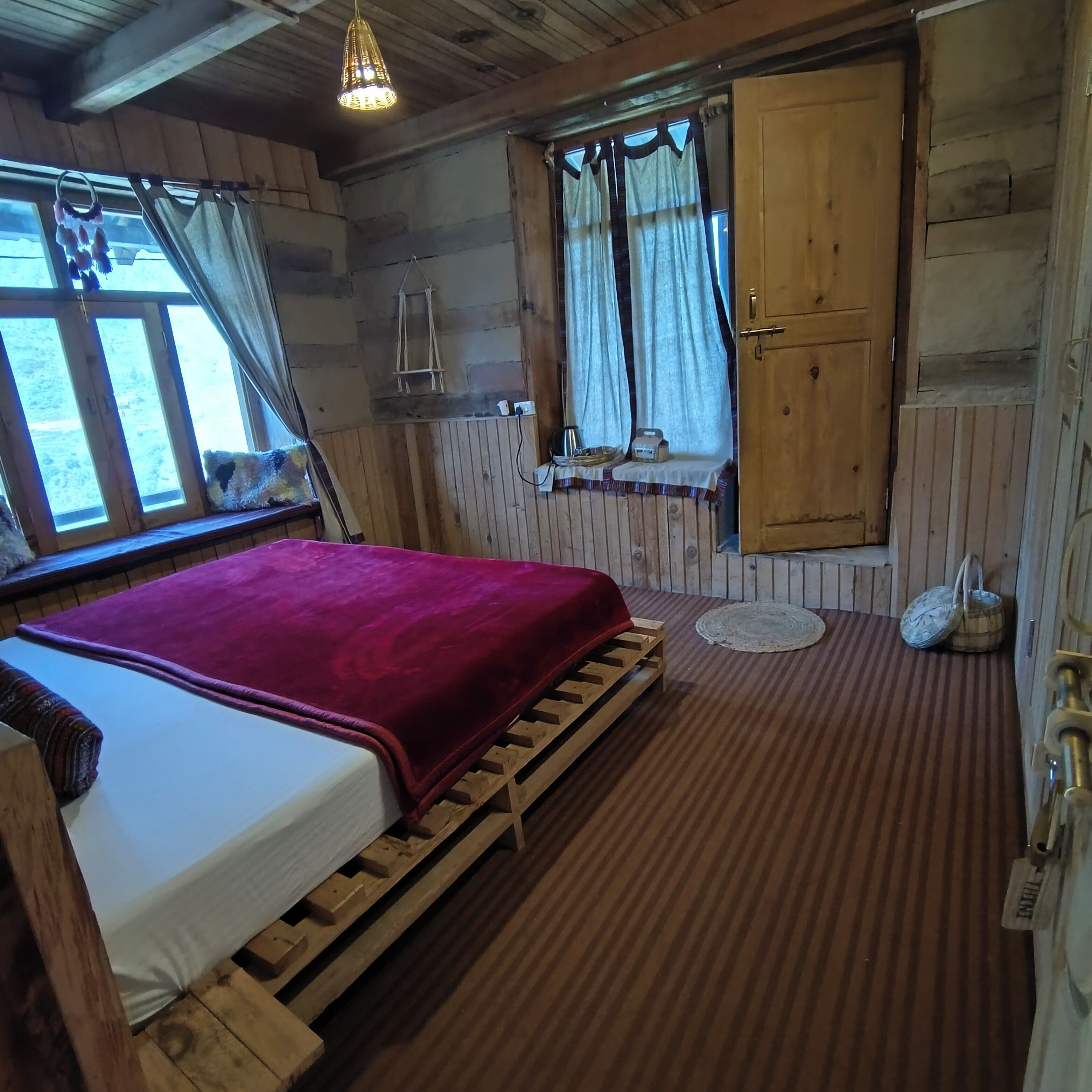 A wooden cabin room with a bed, a window with curtains, a wooden door, and a small table with a kettle and a box.