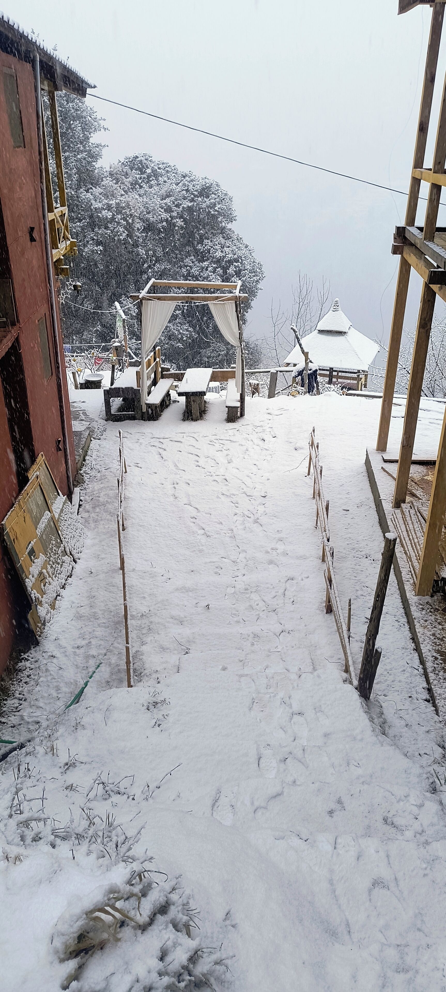 A snow-covered outdoor area with wooden benches and tables, a wooden structure, and a building with a red facade.