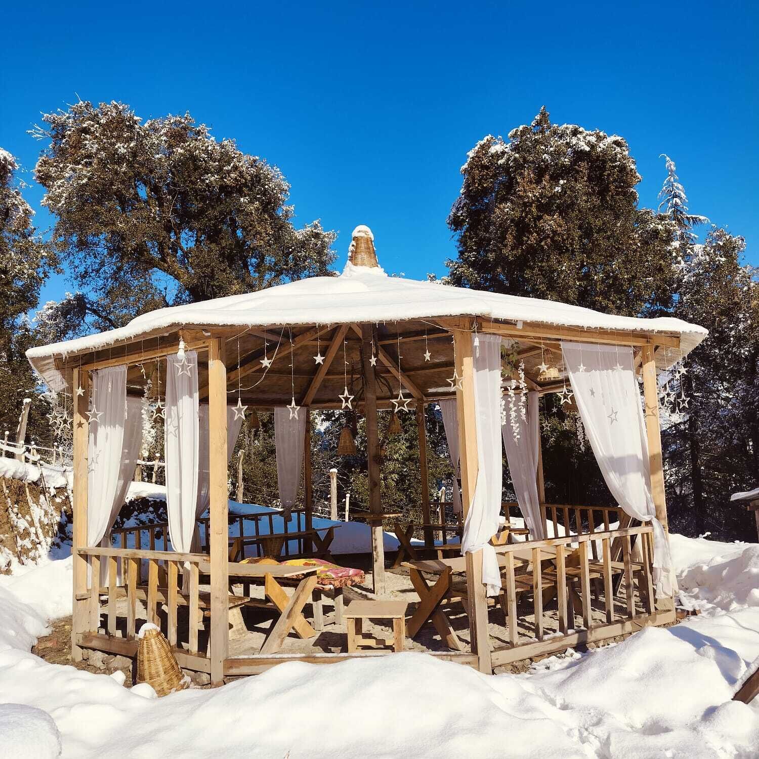 A wooden gazebo with a snow-covered roof, surrounded by trees and snow.