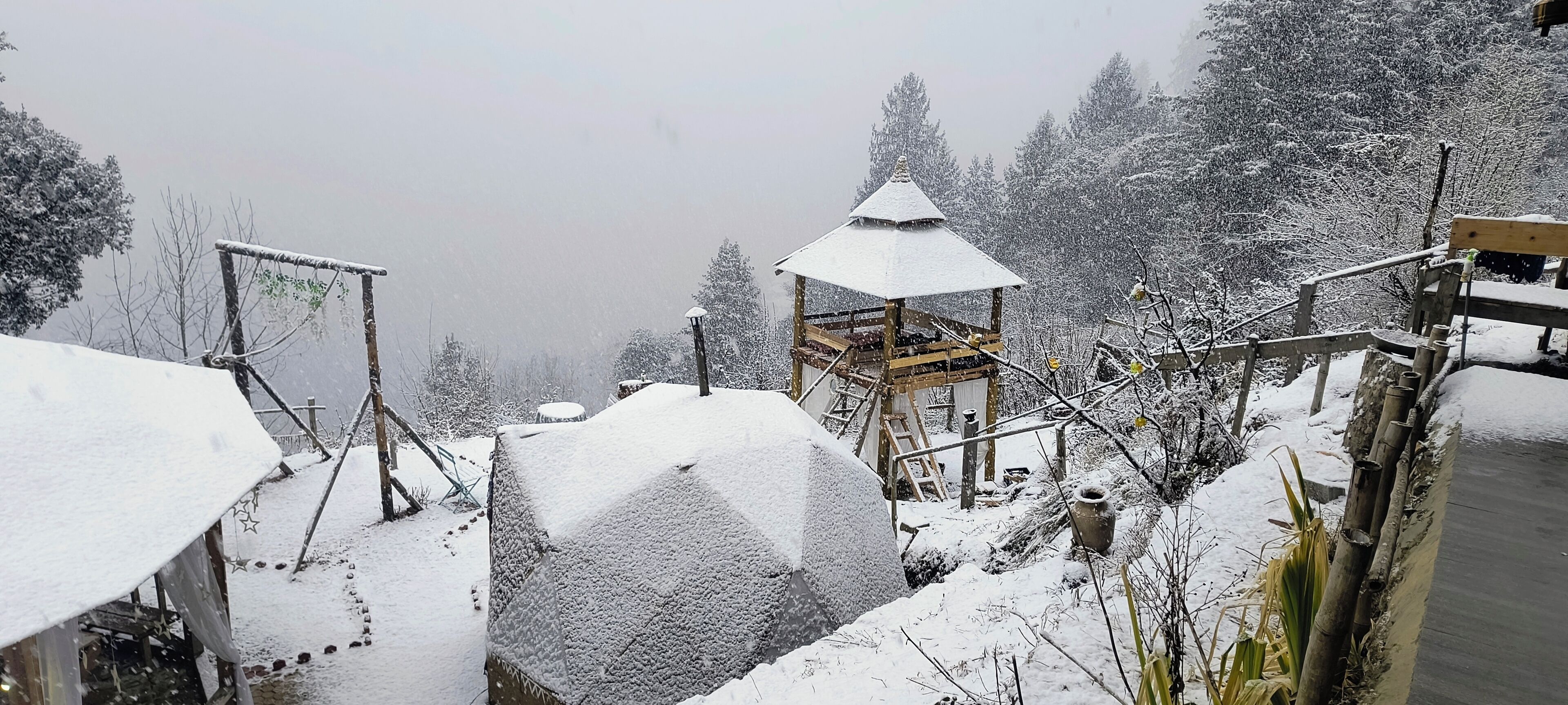 Een besneeuwde buitenruimte met een houten constructie, mogelijk een prieel, omringd door met sneeuw bedekte bomen en een pad.