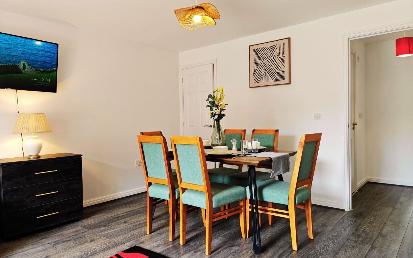 A dining area with a table set for four, surrounded by chairs with green upholstery. A television is mounted on the wall, and a vase with flowers is on the table.