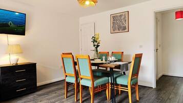 A dining area with a table set for four, surrounded by chairs with green upholstery. A television is mounted on the wall, and a vase with flowers is on the table.