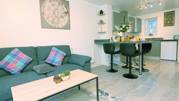 A modern living room with a green sofa, two bar stools, and a coffee table. A kitchen is visible in the background with a dining area. - Barn Fold House with Parking, Garden, Wi-Fi (Telford and Wrekin)