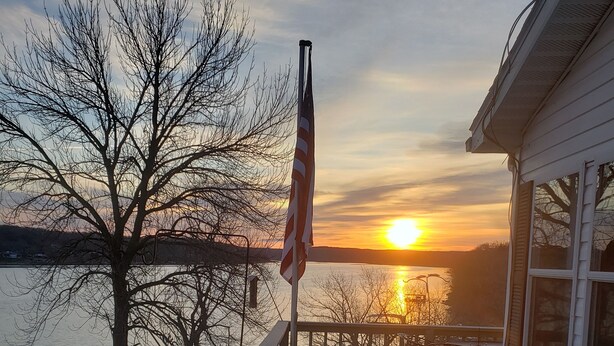 A wooden deck with a railing, overlooking a body of water and a sunset. - Beautiful view on lake! Enjoy 5bdrm 3 bath with 2 decks firepit and private dock (Edwards)
