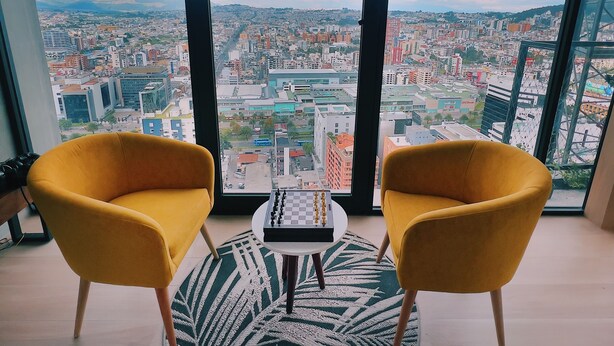 Een kamer met twee gele stoelen, een kleine tafel en een stadsgezicht door grote ramen.