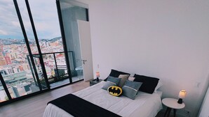 Een moderne slaapkamer met een groot bed, een Batman-thema kussen, een nachtkastje met een lamp en een stadszicht door grote ramen.