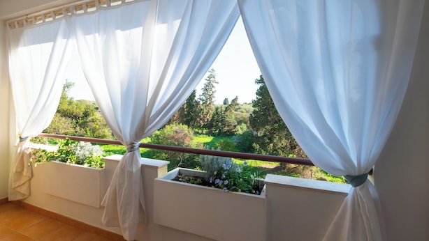 Ein Balkon mit weißen, zurückgebundenen Vorhängen, der einen Gartenblick freigibt.
