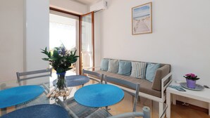 Ein modernes Wohnzimmer mit einer Couch, einem Esstisch mit blauen Platzsets, einer Vase mit Blumen und einem Strandbild an der Wand.