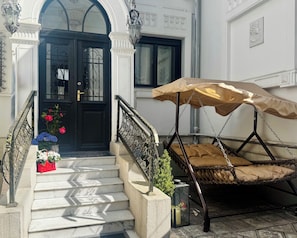 Una escalera conduce a una puerta negra con barandilla de hierro forjado, una silla colgante de mimbre con dosel color beige y plantas en macetas en los escalones.