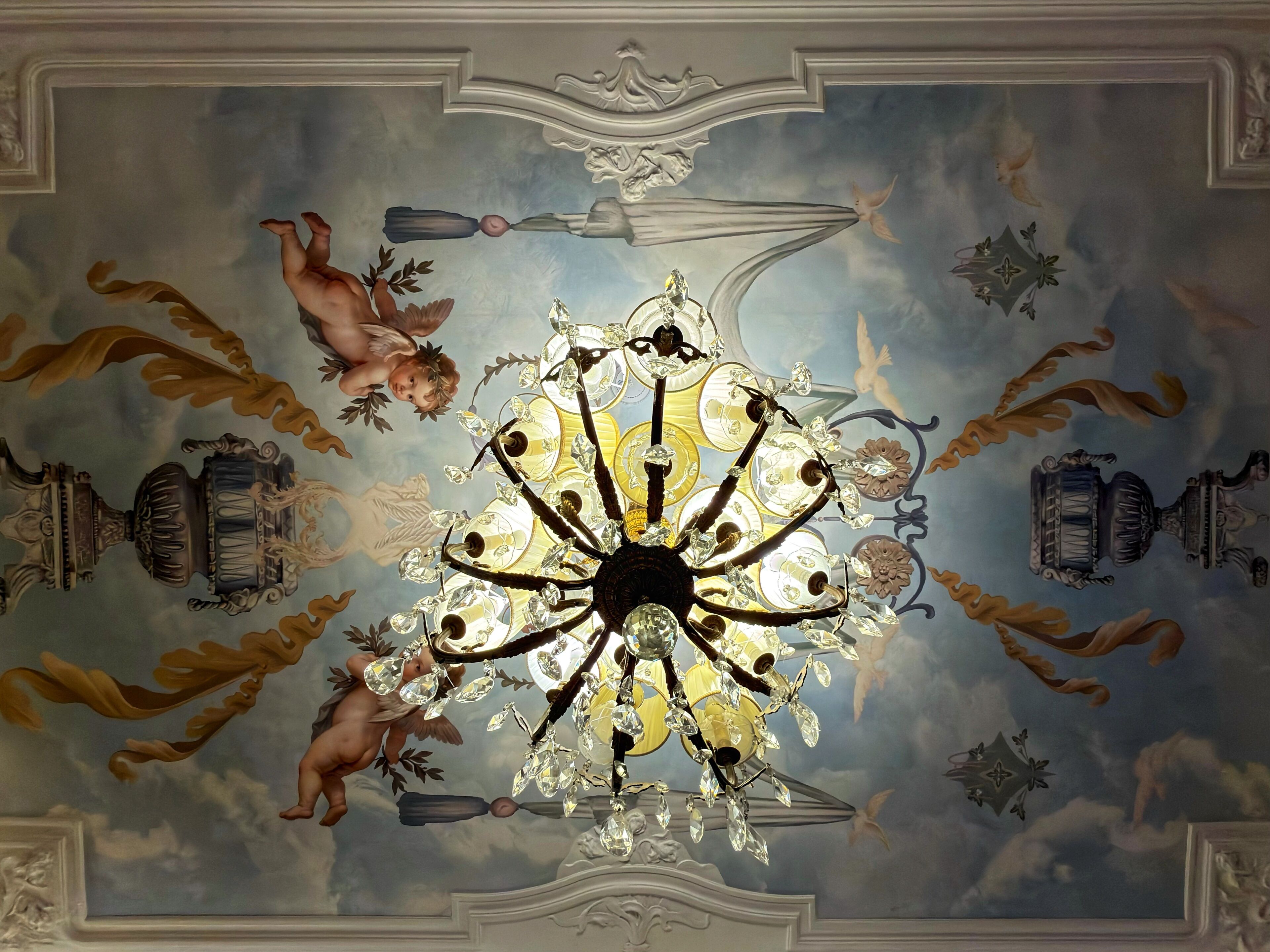 Un plafond orné d’un lustre et d’éléments décoratifs, notamment des putins et des motifs classiques.