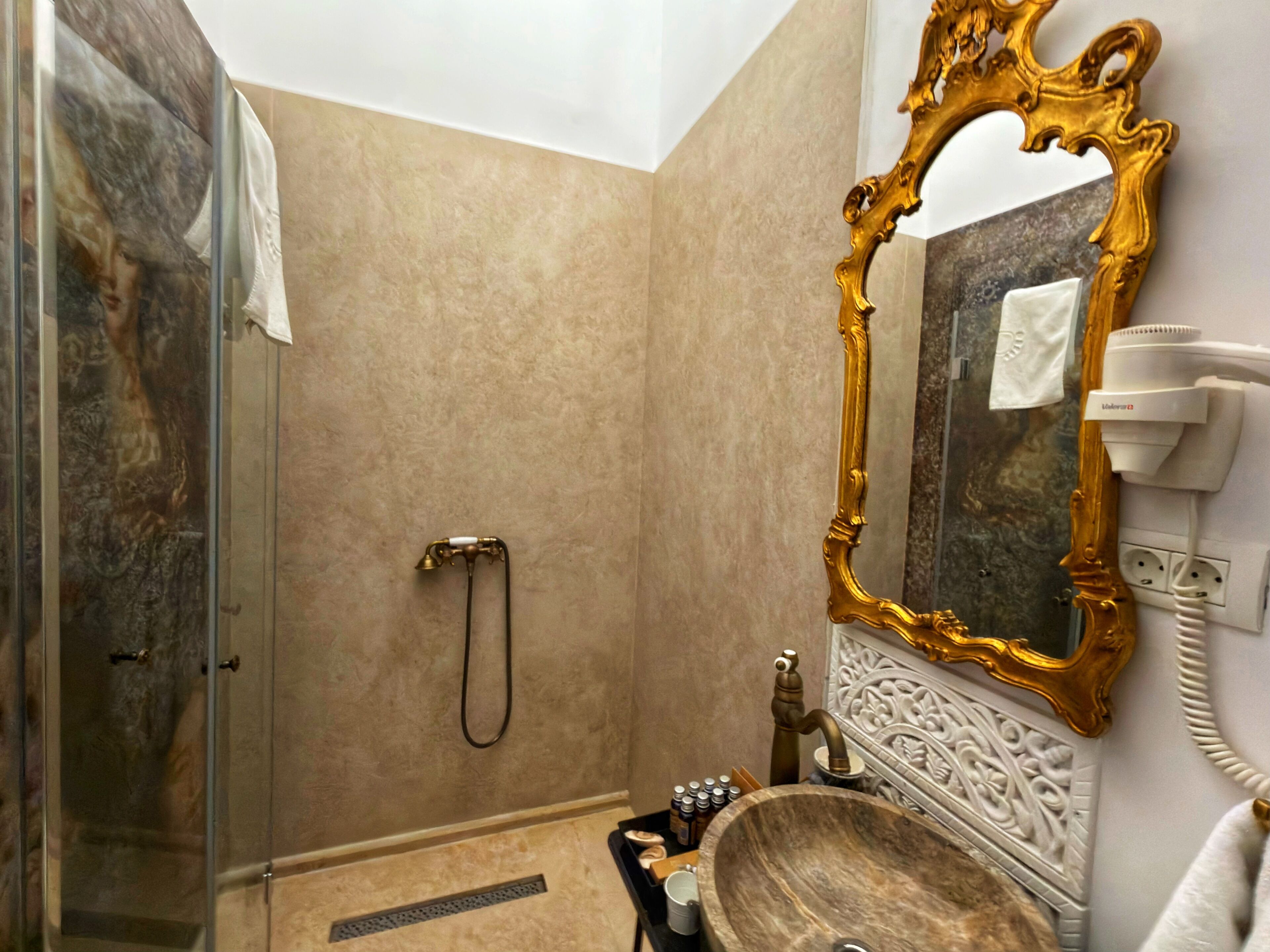 Un baño con pared de mármol, espejo con marco dorado, lavamanos con cuenca de madera y ducha de mano.