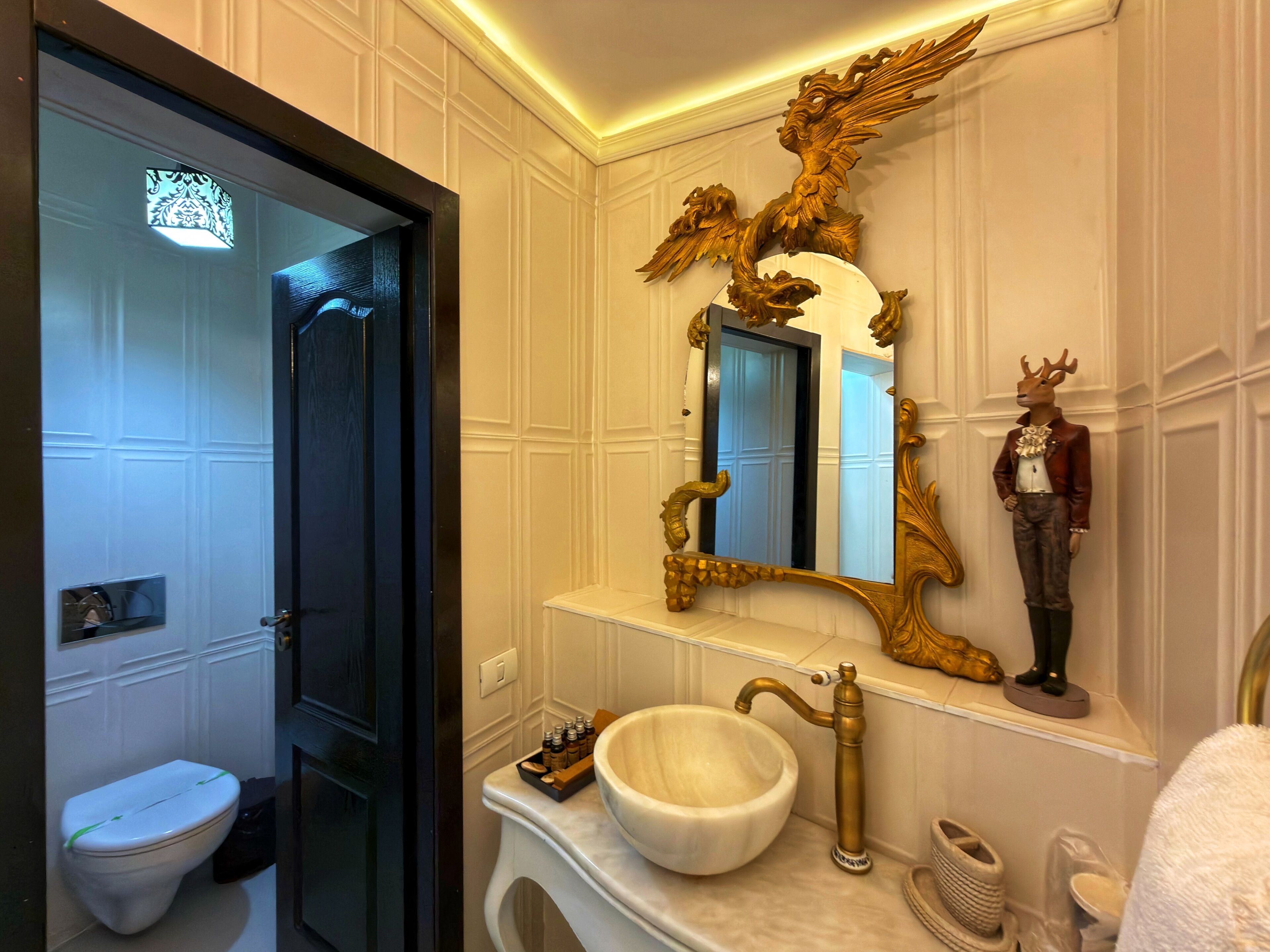 Un baño con inodoro blanco, tocador con lavamanos blanco, espejo dorado ornamentado y una estatua de reno.