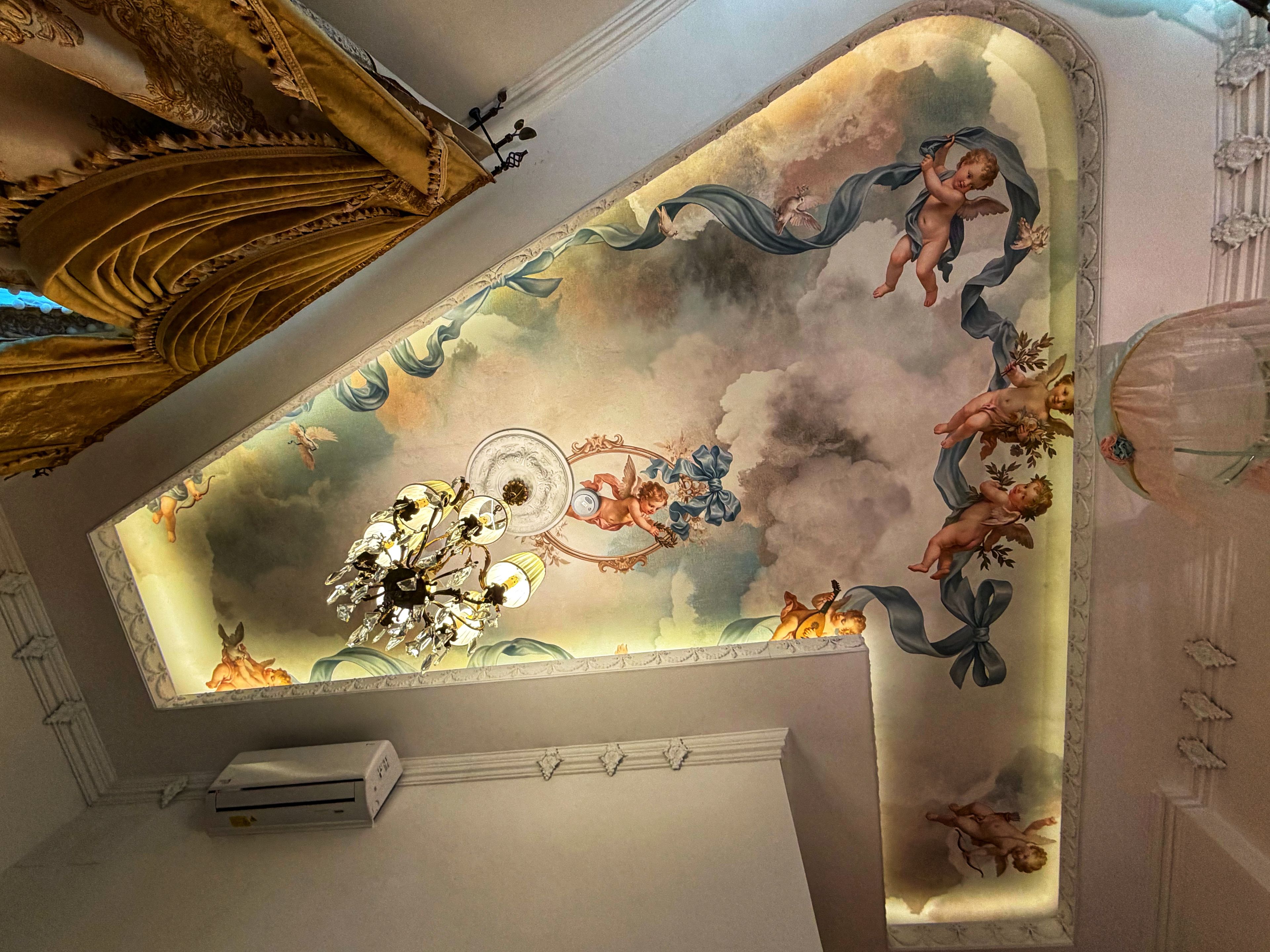 Un techo con frescos ornamentados, una lámpara de araña y un equipo de aire acondicionado instalado en la pared.
