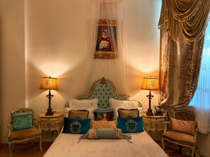 Un dormitorio con cama con dosel, un cabecero ornamentado y un dosel. Hay dos mesitas de noche con lámparas, una silla y una cama con varios almohadones.