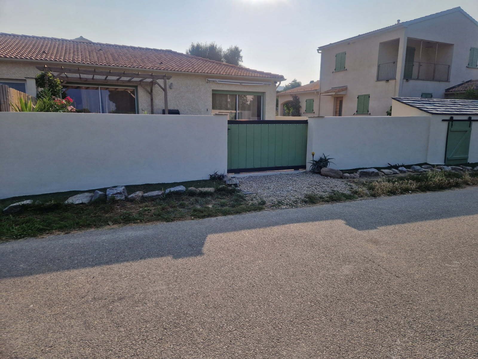 A residential area with two houses, one with a tiled roof and the other with a flat roof. There is a paved driveway and a green gate between the houses.