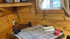 Acogedora habitación tipo cabaña con televisión en un estante, una ventana con cortinas, una mesa con camino de mesa y un libro de visitas.