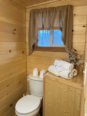 A wooden cabin bathroom with a toilet, a window with a curtain, and a wooden cabinet with towels.