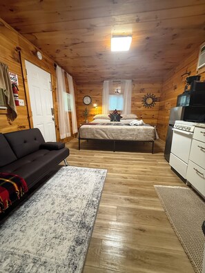 A cozy cabin-style room with a bed, a couch, a kitchenette, and a wooden ceiling. - Riverfront King Cabin,  Cozy RRG Escape for 2 (Stanton)