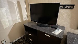 Habitación de hotel con televisor de pantalla plana sobre un mueble de madera oscura, alfombra estampada y una mesita con una silla.