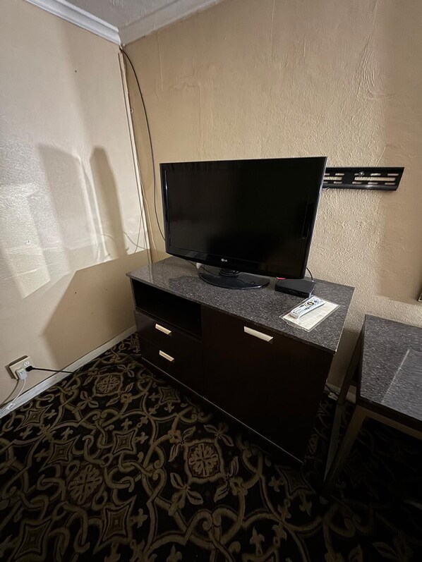 Une chambre d’hôtel avec un téléviseur à écran plat posé sur un meuble en bois foncé, une moquette à motifs et une petite table avec une chaise.