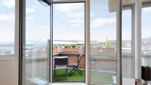 A room with a sliding glass door leading to a balcony with a small table and chairs, a view of the cityscape, and a brick wall with potted plants.