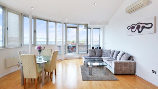 A modern living room with a glass coffee table, grey sofa, and dining area with chairs.
