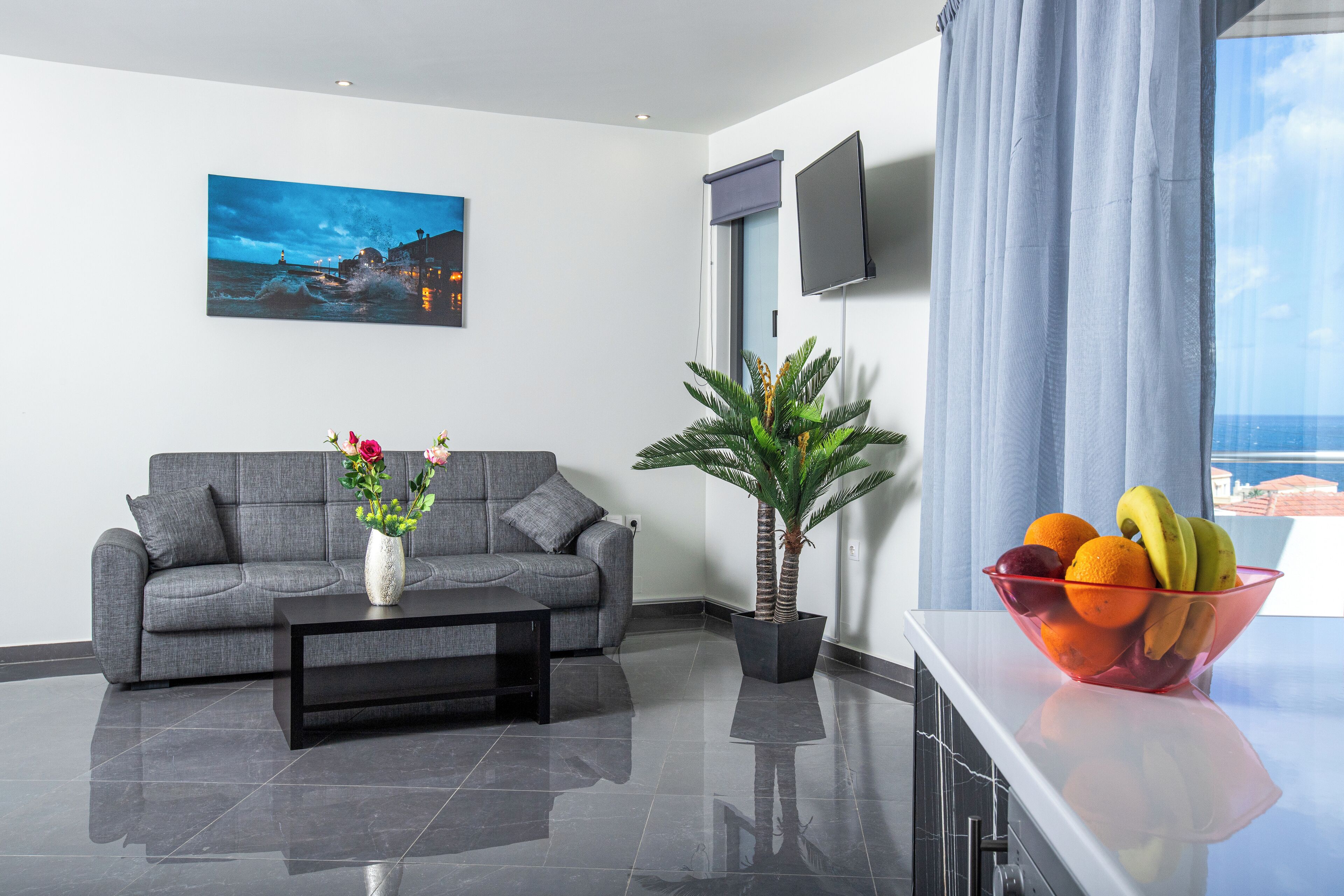 A modern living room with a grey sofa, a black coffee table, a potted plant, and a fruit bowl on a white countertop.