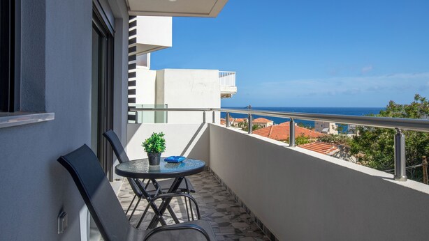 A balcony with a table and chairs, a potted plant, and a view of the sea and buildings. - Mariam Luxury Apts B2 (Chania)