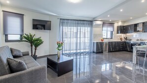 A modern living room with a grey sofa, a coffee table, and a TV mounted on the wall. There is a kitchen area with a black countertop and a dining area with a chair. - Mariam Luxury Apts B2 (Chania)