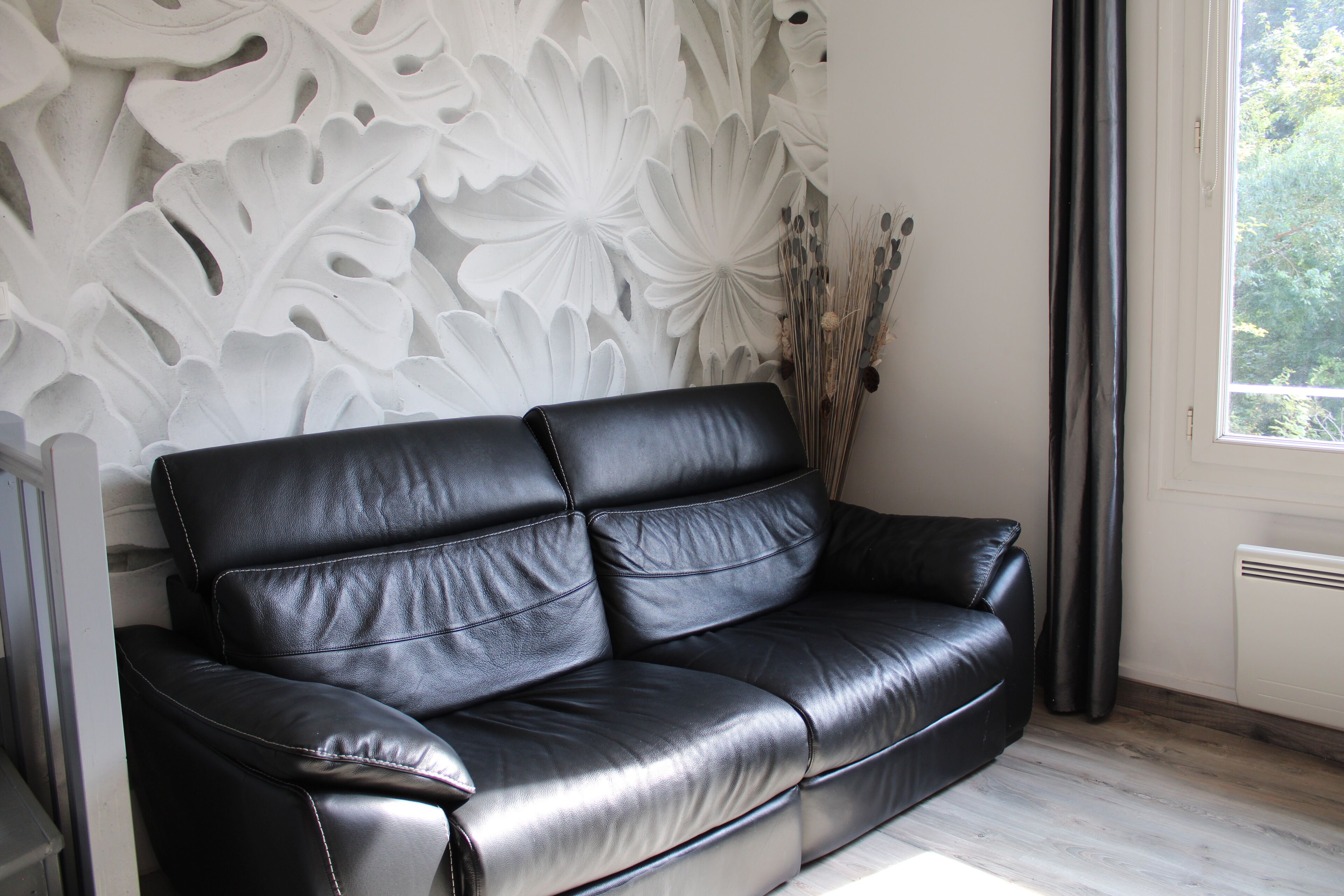 A black leather sofa against a wall with a white floral pattern, a window with a view of trees, and a radiator.
