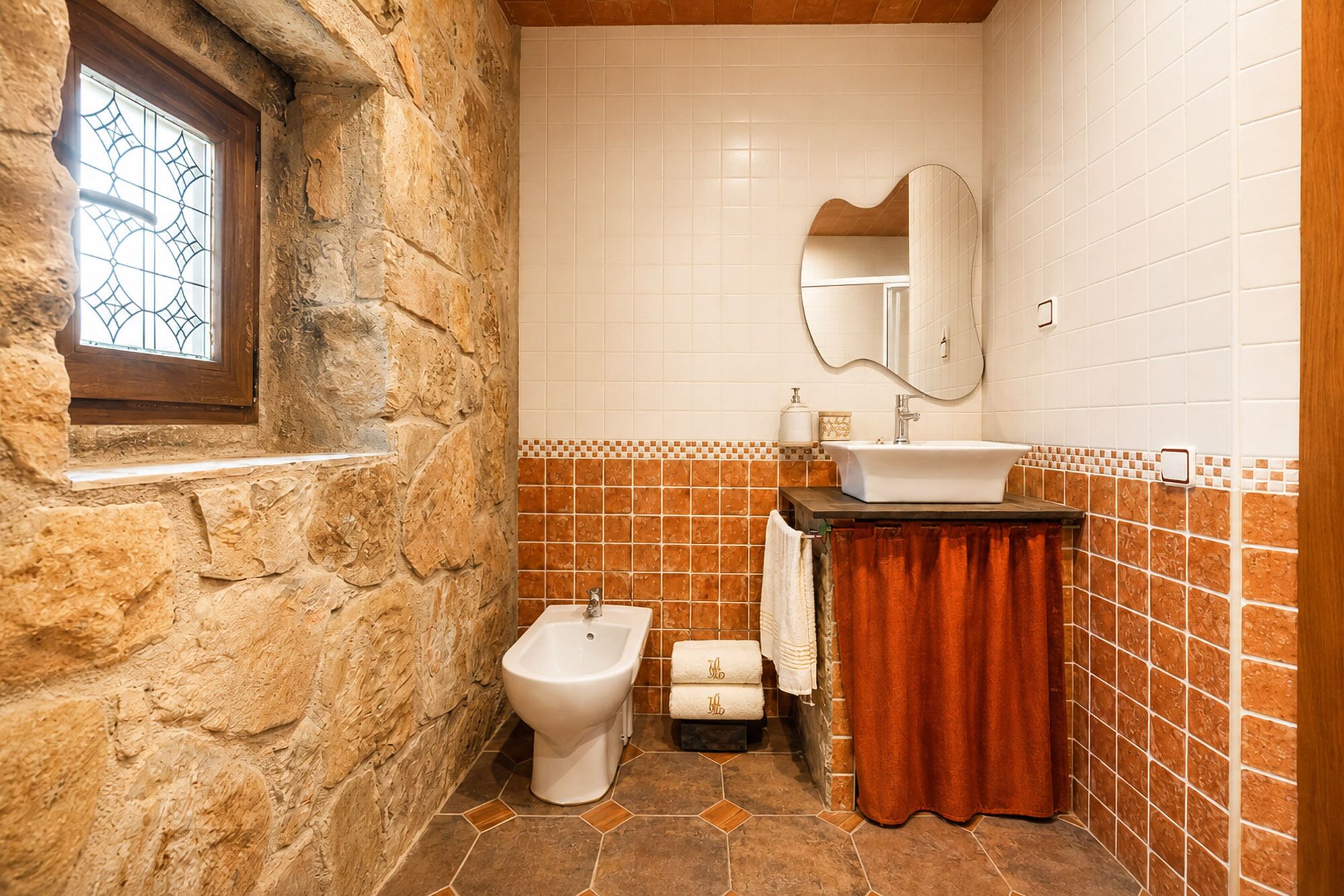 Un baño con pared de piedra, un lavamanos blanco, un bidé y una ventana con diseño decorativo en el vidrio.
