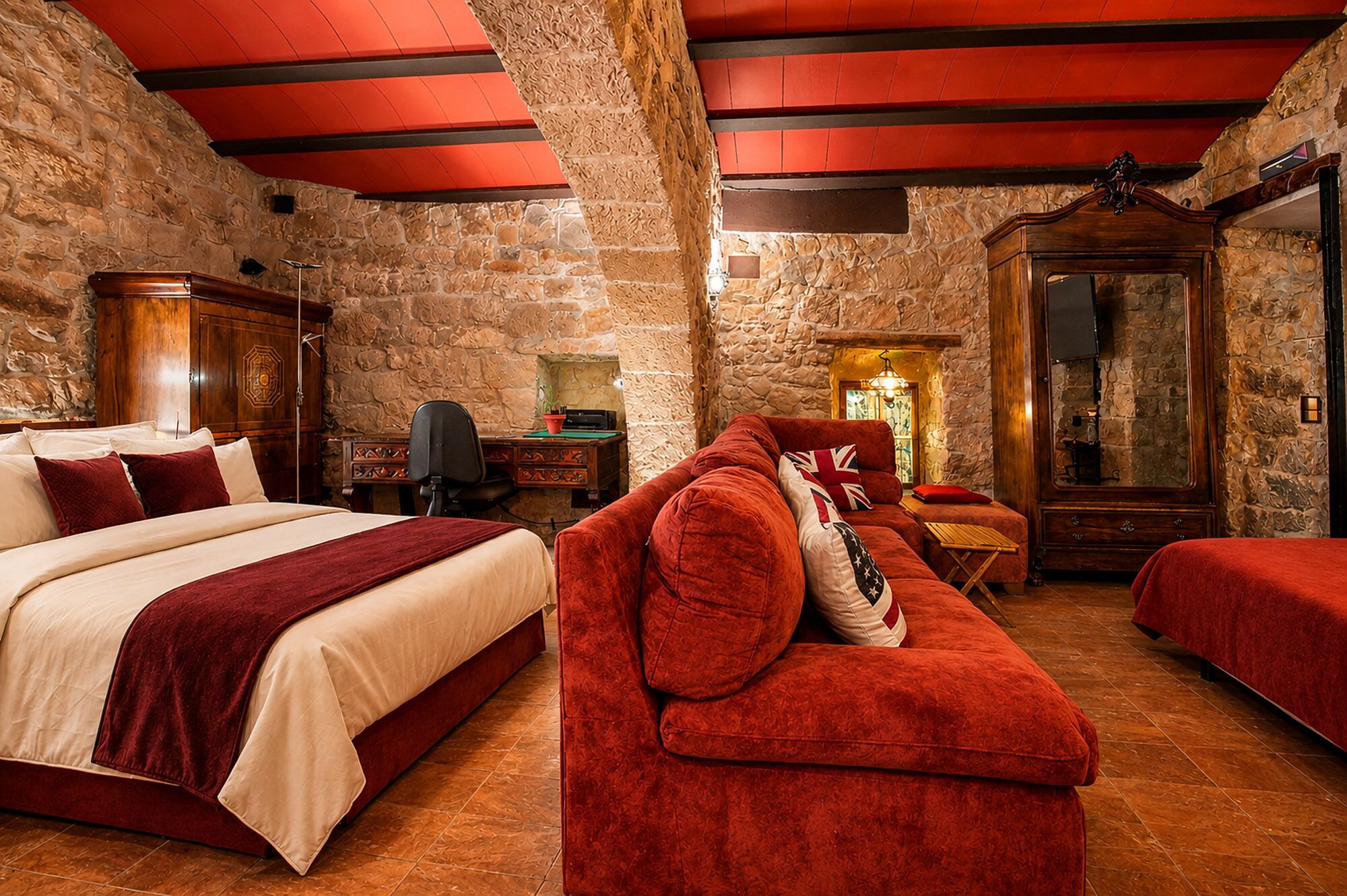 Ein Zimmer mit einem Bett, einem roten Sofa, einem Holzkasten und einer Steinwand.