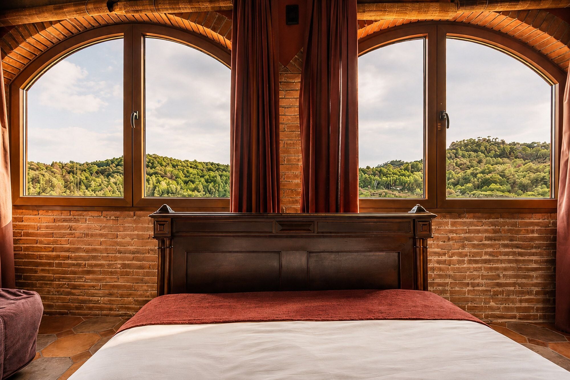 Un dormitorio con una cama grande, dos ventanas con cortinas y vistas a una colina boscosa.