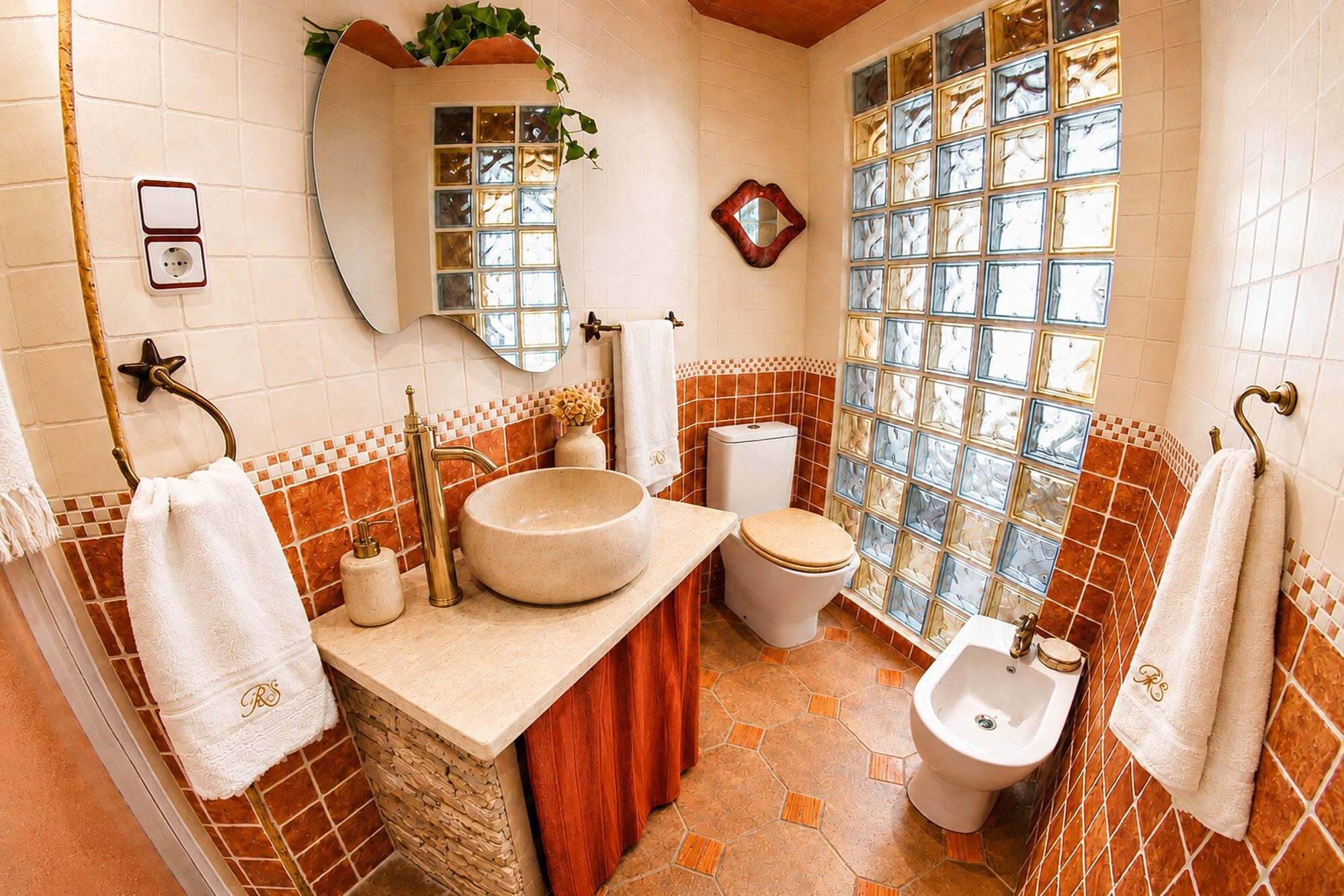 A bathroom with a large mirror, a round sink, a toilet, a bidet, and a window with decorative glass panels.