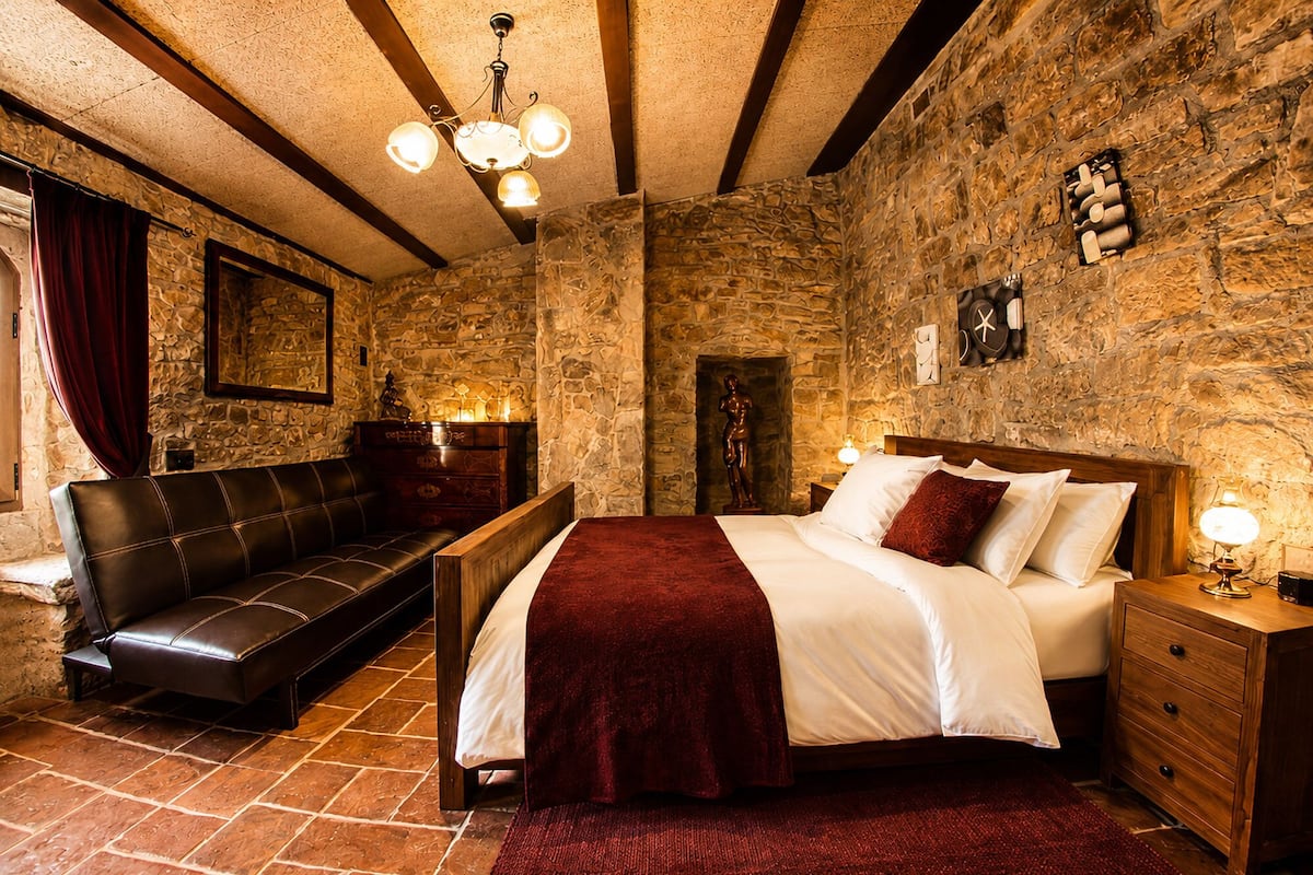 Ein Zimmer mit einer Steinwand, einem hölzernen Bett mit weißer Bettwäsche, einem Ledersofa und einer Kristallleuchtre.