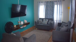 Un salón con paredes color turquesa, un televisor de pantalla plana montado en la pared, un sofá gris con cojines estampados y un suelo de madera.