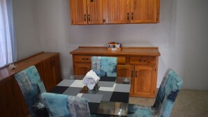 Un comedor con una mesa a cuadros, sillas con diseños azules y blancos, armarios de madera y una ventana con cortinas.