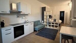 Una cocina moderna con armarios blancos, un horno empotrado y un sofá gris en una sala de estar con alfombra oscura.