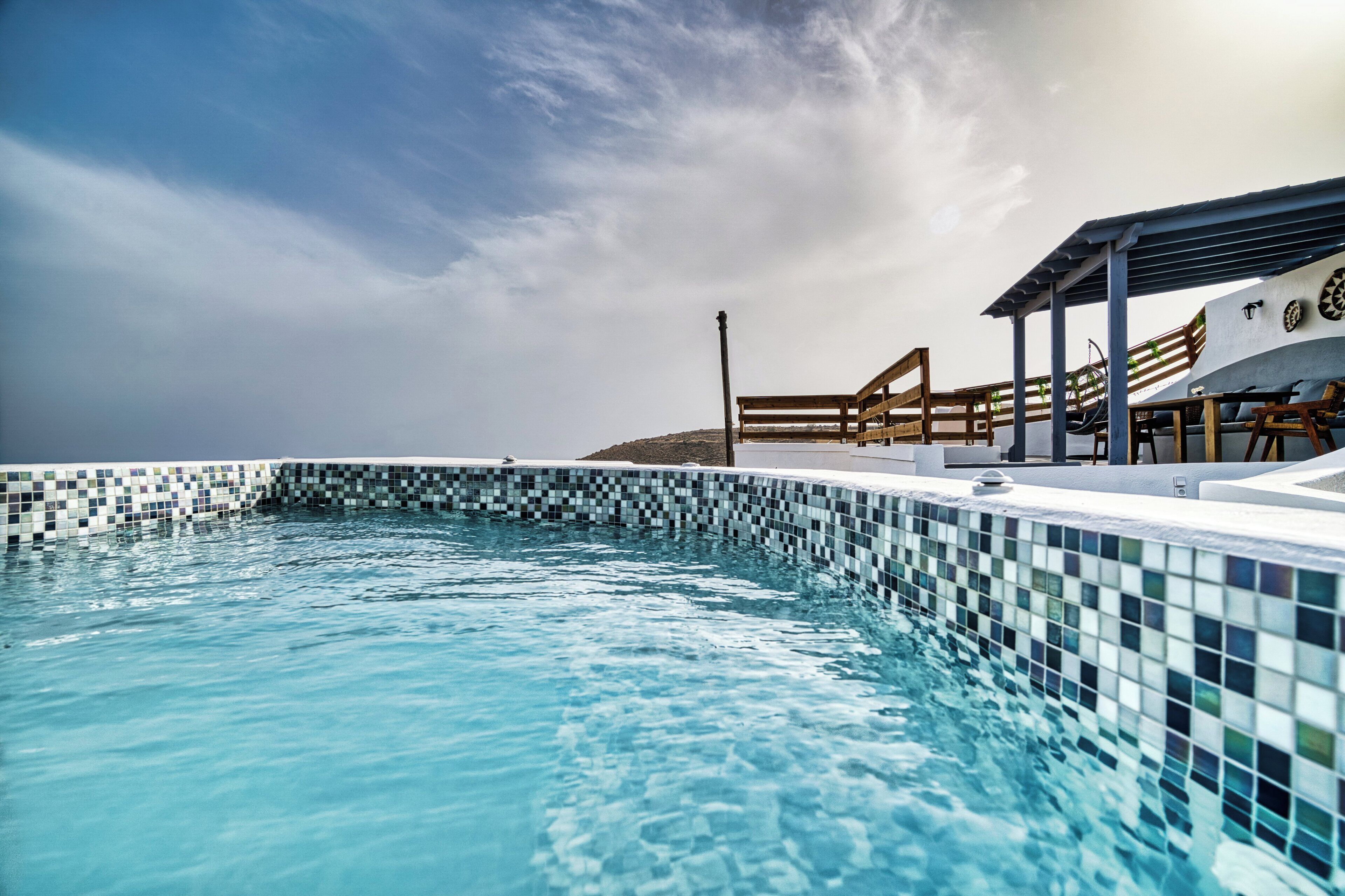 Una piscina con bordes de mosaicos, sillas de madera y un área de descanso cubierta con vista al mar.