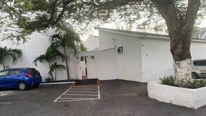 A parking area with a white building, a blue car, and a tree. - Affordable Queen Room w/ Patio | Near Baha Mar & Airport (Nassau)