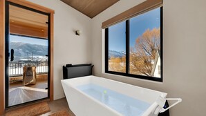 Une salle de bain moderne avec une grande baignoire, une vue sur les montagnes et les arbres par une fenêtre, et une porte donnant sur un balcon.