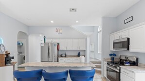 A modern kitchen with white cabinets, a central island with blue bar stools, stainless steel appliances, and a marble countertop.