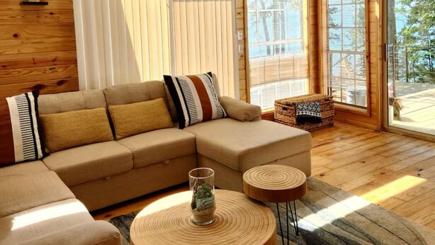 Un interno moderno di una baita con un divano componibile, tavolini in legno rotondi e ampie finestre che offrono luce naturale e vista sulla natura.
