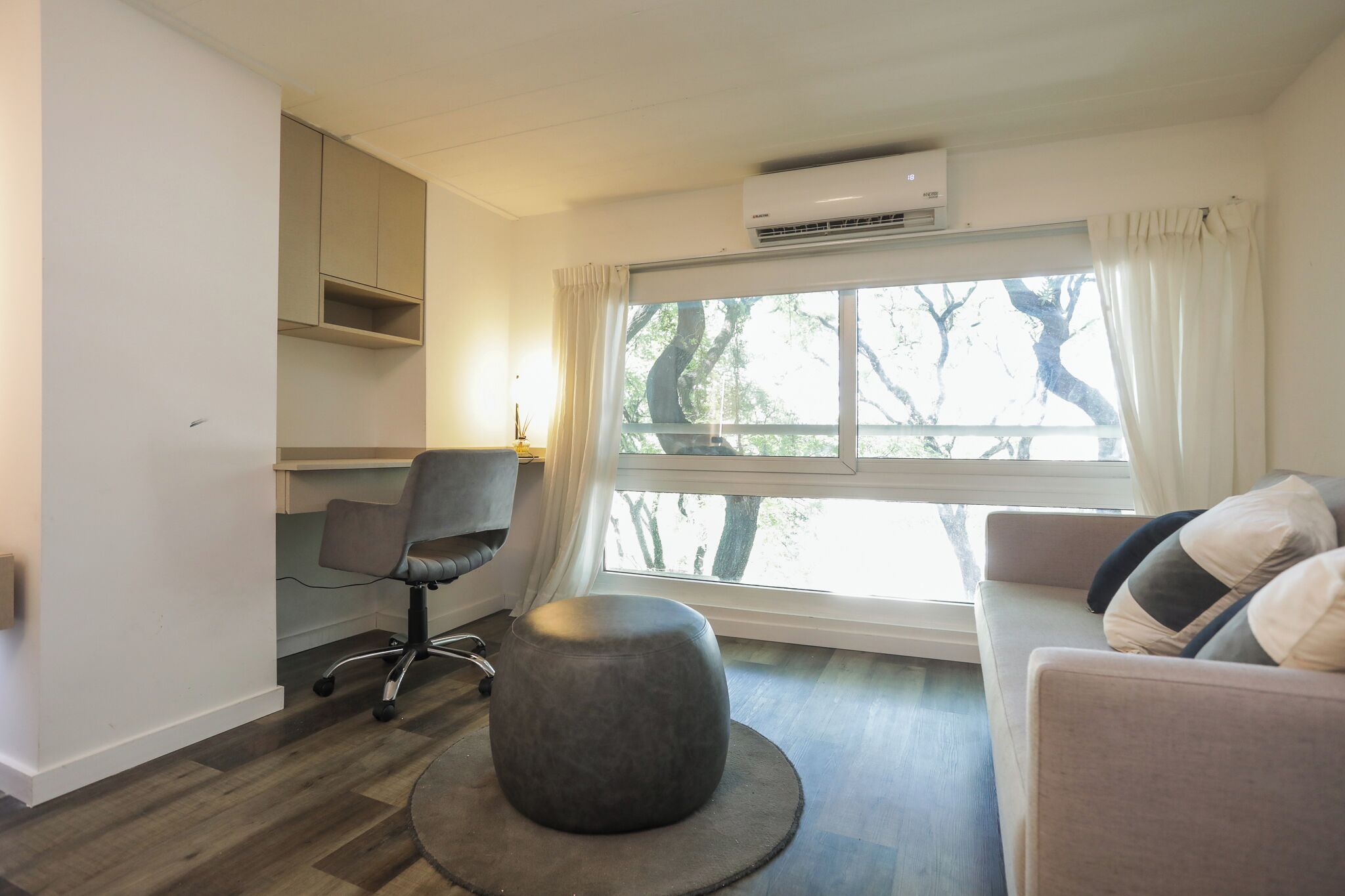Ein modernes Wohnzimmer mit einem grauen Ottomanen, einem grauen Bürostuhl, einem beigefarbenen Sofa mit Kissen, einem Fenster mit Vorhängen und einer Klimaanlage.