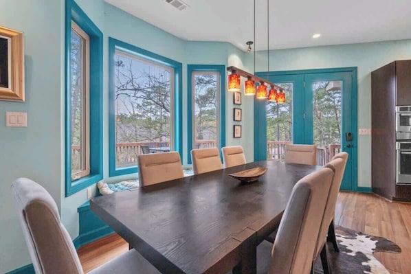A dining room with a large table and chairs, a kitchen with a stove, and large windows with a view.