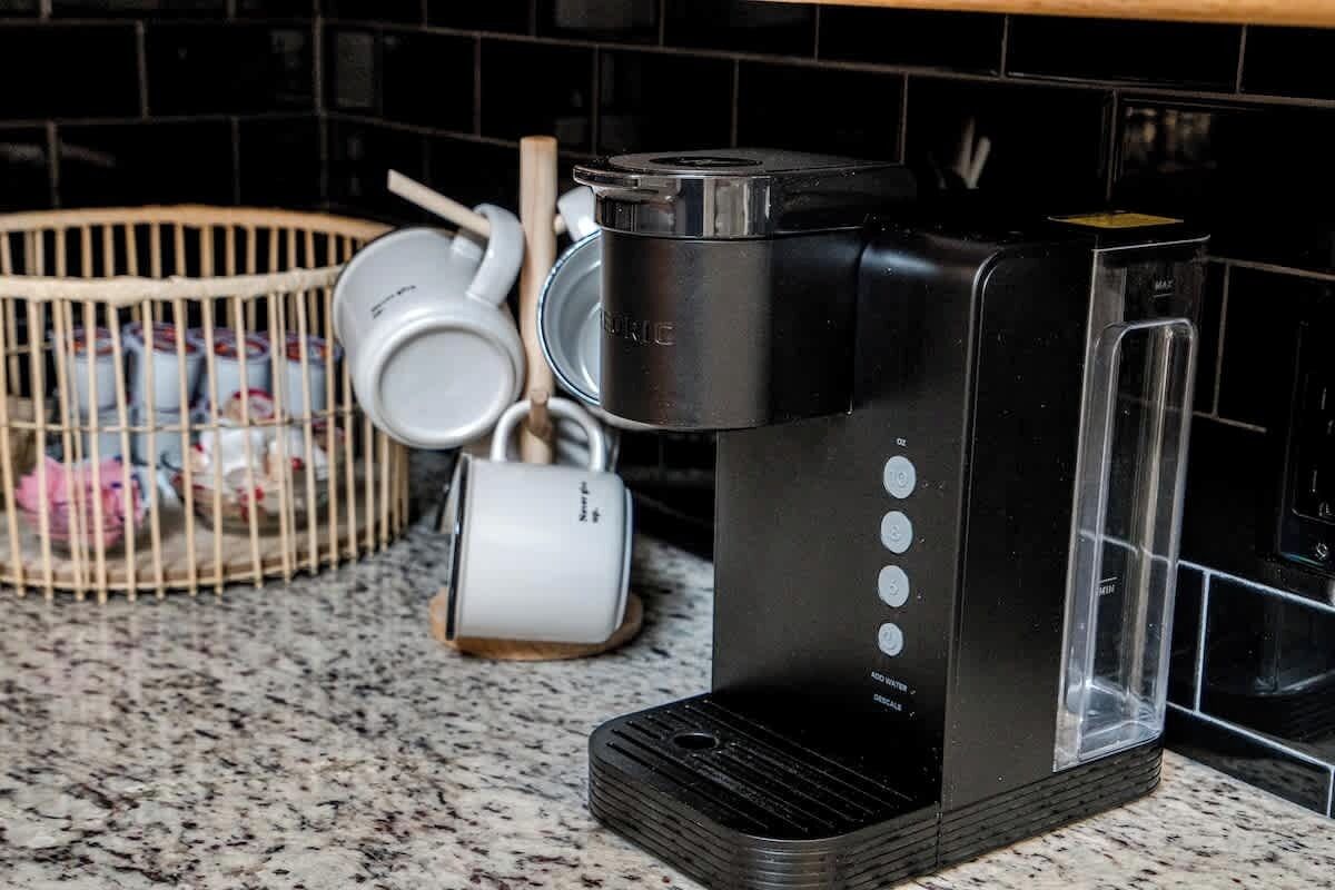 A coffee maker on a kitchen counter with a basket of cups and a wooden spoon.