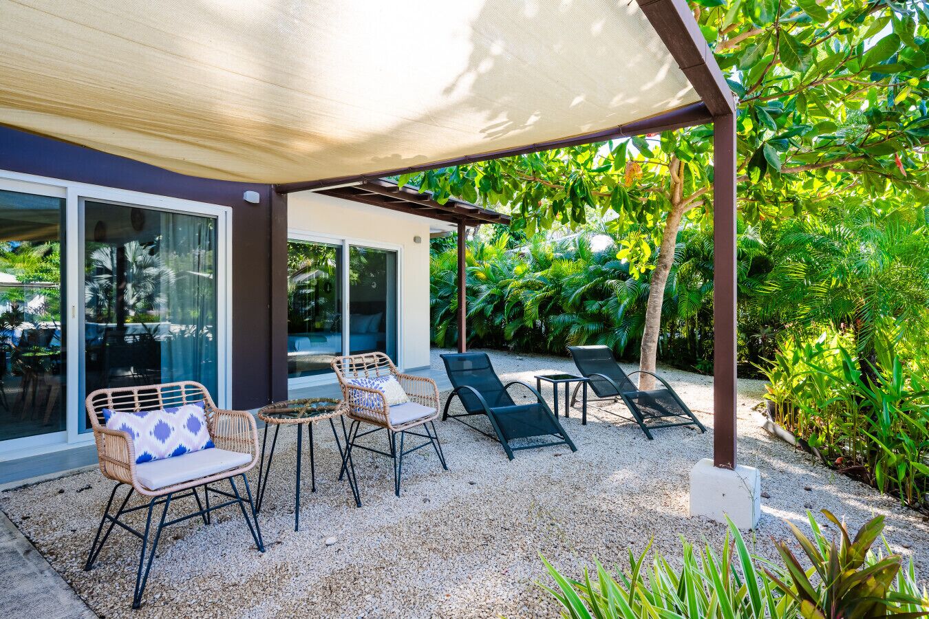An outdoor seating area with wicker chairs and lounge chairs, a pergola providing shade, and a view of a lush garden.