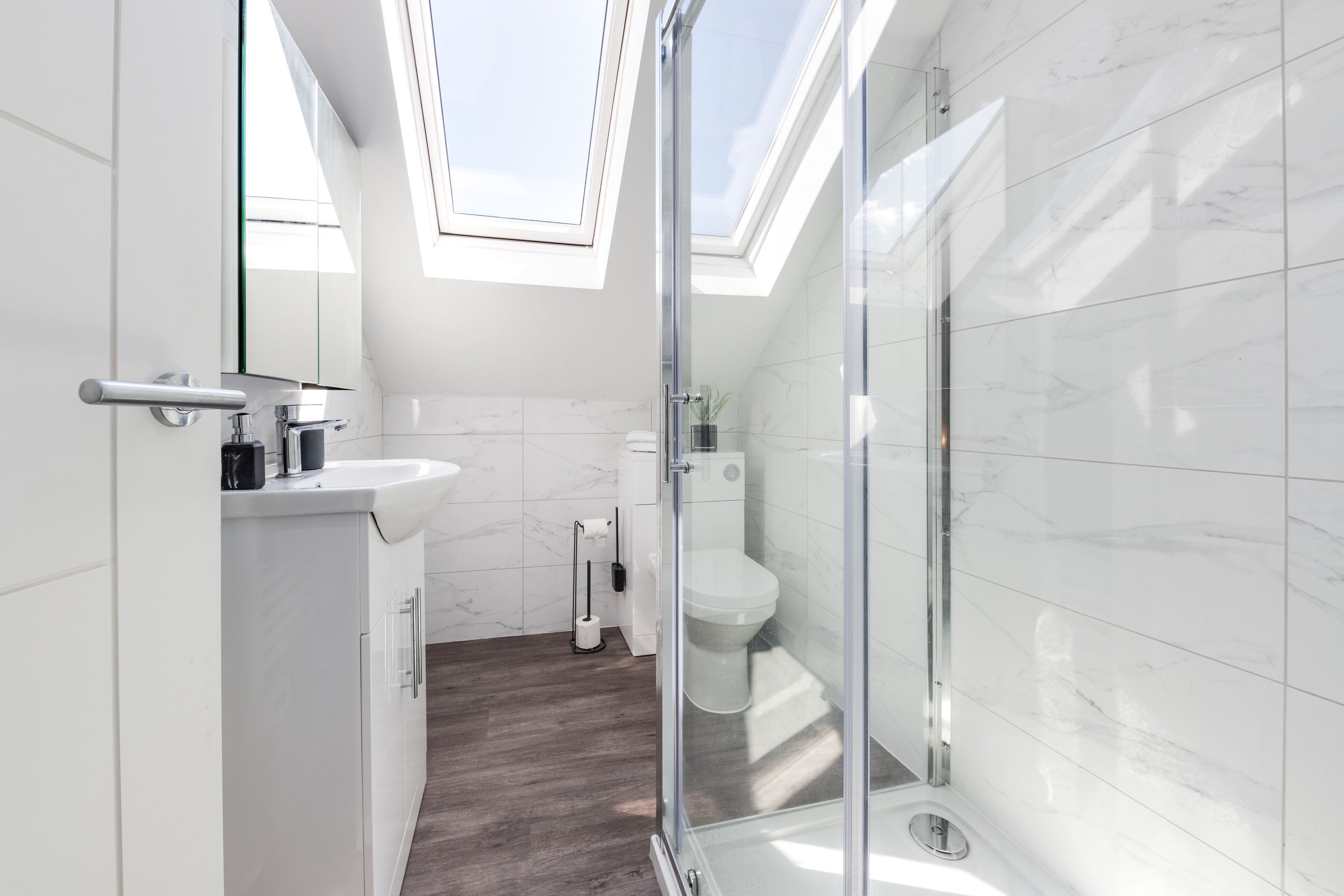 A modern bathroom with a skylight, white marble tiles, a glass shower enclosure, and a wooden floor.